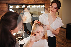 Hairdresser and make up artist work together in beauty room. Young blonde woamn sit on chair with clossed eyes. Applying