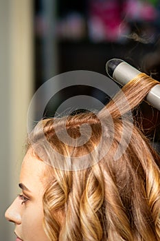 Hairdresser makes curls with a curling iron for the young woman with long brown hair in a beauty salon.