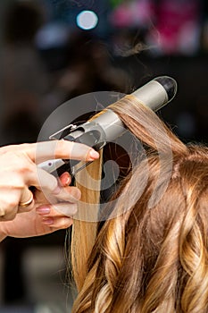 Hairdresser makes curls with a curling iron for the young woman with long brown hair in a beauty salon.