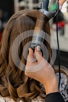 The hairdresser prepares a woman, makes a hairstyle with curls using a curling iron. Long light brown natural hair. Self