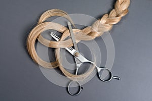 Hairdresser professional thinning scissors or shears with braid strand of blonde hair on grey background. Beauty salon. Hair