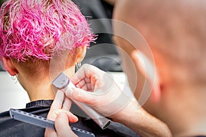 Hairdresser shaving nape and neck with electric trimmer of a young caucasian woman with short pink hair in a beauty