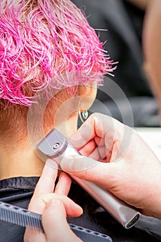 Hairdresser shaving nape and neck with electric trimmer of a young caucasian woman with short pink hair in a beauty