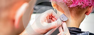 Hairdresser shaving nape and neck with electric trimmer of a young caucasian woman with short pink hair in a beauty