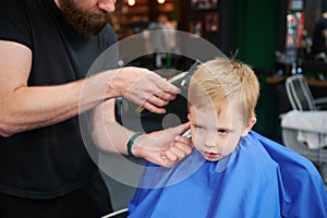 Hairdresser using electric shaver to cut boy's hair. Little kid getting first haircut in barbershop.