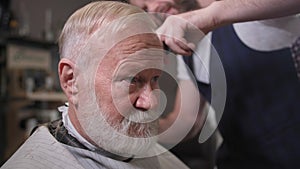 hairdressing salon, professional barber cutting hair with scissors and comb to stylish elderly man in barbershop, close