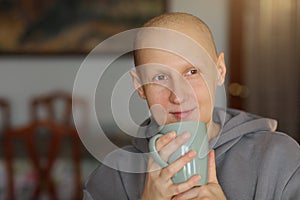 Hairless happy woman is holding cup of hot drink, coffee or tea. Bald female is having remission after cancer disease