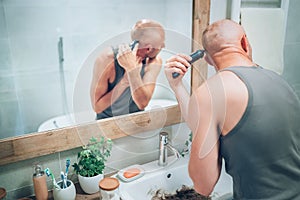 Hairless man ending shaving off all his hairs looking in bathroom mirror using an electric rechargeable Trimmer. Radical decision photo