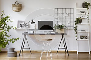 Hairpin stool standing by the wooden desk with mockup computer screen, metal lamp and coffee cup in real photo of white home offic