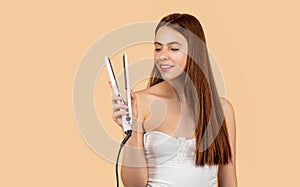 Hairstyle. Beautiful smiling woman ironing long hair with flat iron. Happy woman straightening hair with straightener