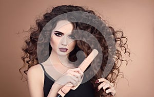 Hairstyling. Curly Beauty hair. Glamour portrait of beautiful woman model holding straightening iron. Brunette with marsala matte