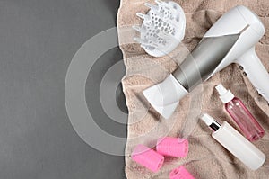 Hairstyling tools (white hairdryer, hairspray, pink curler, spray) on beige towel. Top view