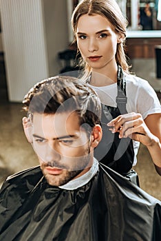 Hairstylist looking at camera while cutting hair to handsome young man