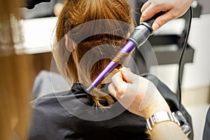 The hairstylist makes curls hairstyle of long brown hair with the curling iron in hairdresser salon, close up.
