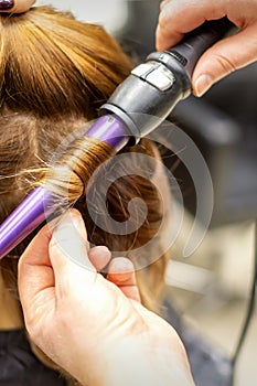 The hairstylist makes curls hairstyle of long brown hair with the curling iron in hairdresser salon, close up.