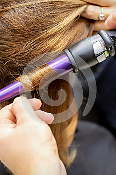 The hairstylist makes curls hairstyle of long brown hair with the curling iron in hairdresser salon, close up.