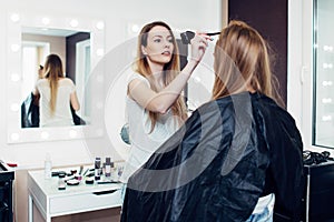 Hairstylist parting the long fair hair of young female customer sitting in cape at hairdressing salon