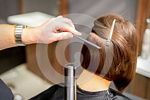 Hairstylist is straightening short hair of young brunette woman with a flat iron in a hairdresser salon, close up.