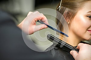 Hairstylist is straightening short hair of young brunette woman with a flat iron in a hairdresser salon, close up.