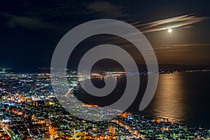 Hakodate City night view from Mt. Hakodate observatory, big bright moon light up the sea, golden reflection on surface