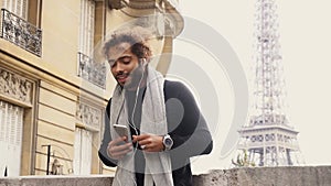 Half African tourist listening to new songs in earphones with smartphone near Eiffel tower.