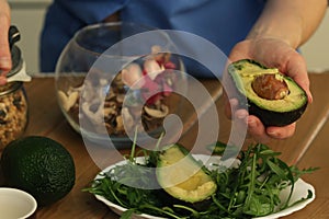 half avocado in human hand and arugula salad in plate with knife close up photo on kitchen table