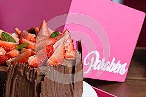 Half a chocolate cake with strawberries, basil leaves, blackberry jam and chocolate plates. Written in Portuguese: Congratulations photo