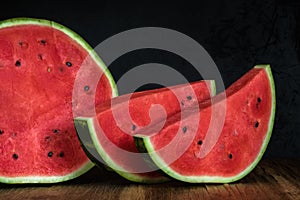 Half of a cut ripe watermelon with two large slices and red juicy pulp on a wooden table against a dark background of black