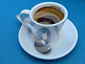 Half empty full white porcelain coffee cup, saucer and a tea spoon on blue background.