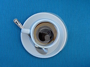 Half empty full white porcelain coffee cup, saucer and a tea spoon on blue background. Top view.