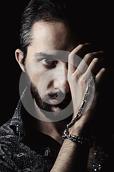 Only half of the face is lit. The hand covers half the face. On the hand is a silver chain. Man portrait