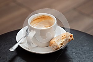 Half full cup of espresso macchiato with metal spoon and half eaten vanilla macaron on saucer. Black leather table