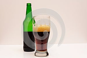 Half full transparent glass with dark beer and half empty green beer bottle on a light background