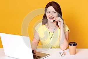 Half lengh photo of woman operator sitting at office desk, looking aside, touching headset,  over yellow background,