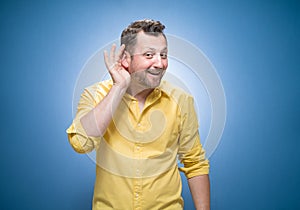 Half length man holding palm near his ear trying to listen something interesting over blue background, dresses in yellow shirt.
