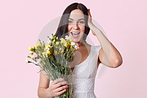 Half length portrait of pretty brunette woman holding bunch of flowers, laughing while looking directly at camera, keeps hand on