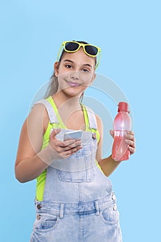 A young brunette girl wearing  denim overalls and green sunglasses shorts stands with a phone and with a bottle of water