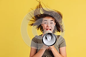 Half-length portrait of young girl, student shouting at megaphone isolated on yellow studio backgroud.
