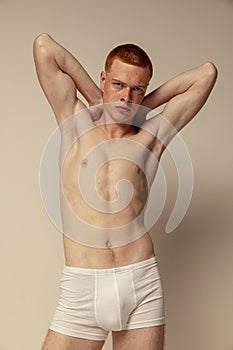 Half-length portrait of young muscular red-haired man posing in white underwear, boxers isolated over grey studio
