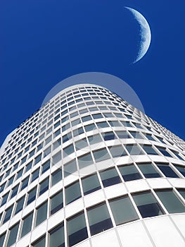 Half a moon over a building skyscraper, shining in bright blue sky. Below the crescent of quarter moon over a tall
