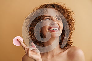 Half-naked ginger woman smiling while showing cosmetic sponge