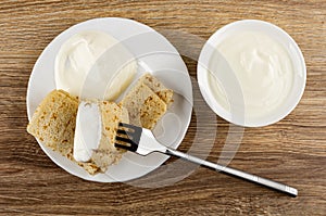 Half of pancake roll with sour cream strung on fork, pancakes in plate, bowl with sour cream on table. Top view