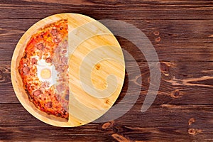 Half a pizza with beef sausages, eggs and bacon on a round wooden platter which is on wood rustic background, top view, copy space