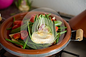 A half of ripe organic green artichoke and vegetables on the clay dish while cooking traditional Moroccan tagine