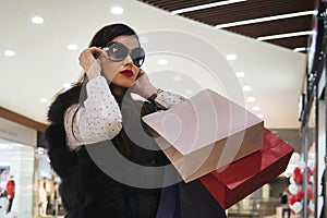 Half-shot of young attractive girl putting on sun glasses after shopping in shopping centre. Trendy brunette girl with paper bags