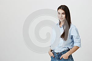 Half-turned cute european businesswoman with hands in pocket, expressing confidence and readiness, standing over gray