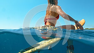 HALF UNDERWATER: Young woman sits on her surfboard and waits for big waves.
