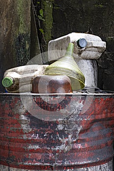 Half view of old and dented red and white oil barrel with empty plastic containers piled on top