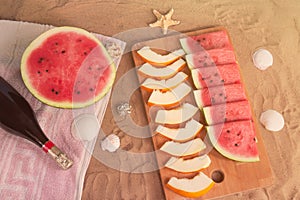 Half of watermelon, slices of watermelon and melon, bottle of champagne, starfishes and seashells on sand beach.