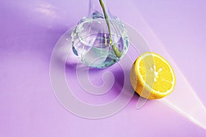 Half of a yellow lemon lies next to a glass vase with water on a lilac background of waters by the glare of the sun